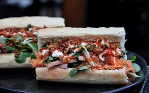 Zwei appetitliche Banh Mi Sandwiches mit Fleisch, Gemüse, knusprigen Zwiebeln und Saucen auf einem Teller.