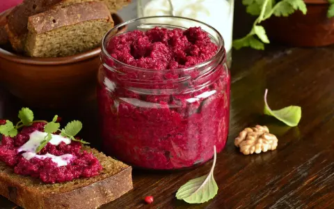 Glas mit leuchtend rotem Rote Bete Pesto, daneben Brot mit Pesto und Joghurt. Walnuss und Kräuter garnieren die Szene.