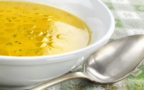 Una ciotola bianca piena di brodo di carne dorato, con goccioline d'olio in superficie. Un cucchiaio d'argento è appoggiato accanto.