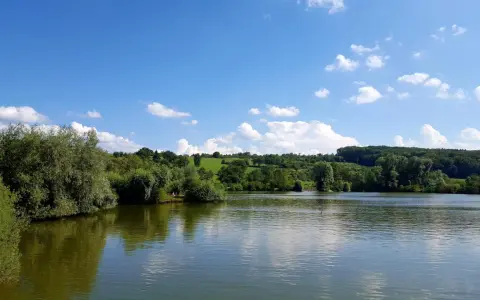 Ein sonniger Tag am Badesee Esslingen. Grüne Hügel, Bäume und ein blauer Himmel mit weißen Wolken spiegeln sich im ruhigen Wasser.