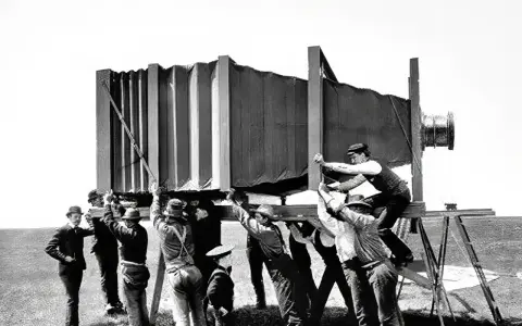 Grupa mężczyzn w historycznych strojach ustawia gigantyczny aparat fotograficzny. Wyobraź sobie, kiedy wynaleziono aparat fotograficzny i jak wielkie musiały być pierwsze urządzenia!