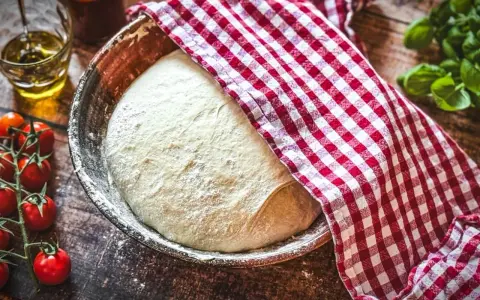 Frischer Pizzateig ruht in einer Schüssel, bedeckt mit einem rot-weiß karierten Tuch. Daneben liegen Kirschtomaten und ein Glas Olivenöl – die perfekten pizzateig zutaten.
