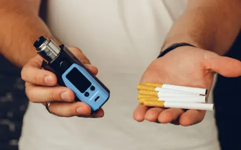 Man holds a vape in one hand and cigarettes in the other, posing the question: que es mejor el vaper o el tabaco.