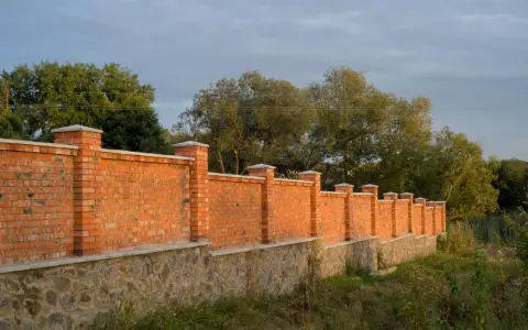 Solidny płot murowany z cegły i kamienia, z ozdobnymi słupkami, otacza teren porośnięty zielenią.