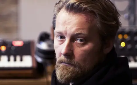 Bartek Dziedzic, z zarostem i lekko zaczerwienionym czołem, siedzi przed syntezatorami.