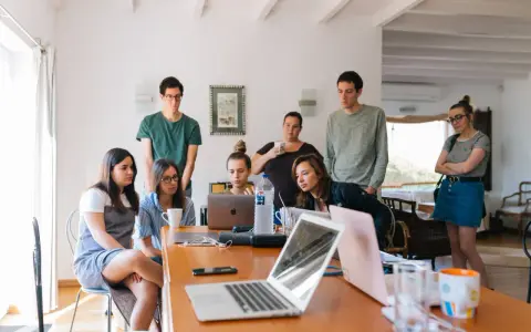 Grupa studentów na którym roku studiów są praktyki, pochylona nad laptopami, dyskutuje przy drewnianym stole.
