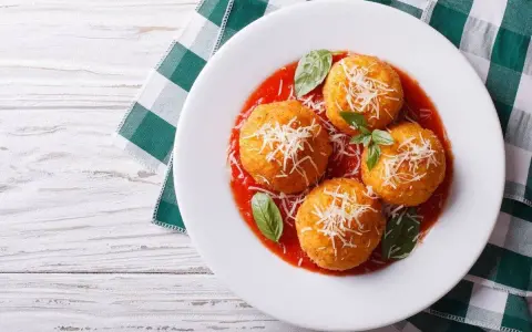 Quattro arancini di riso dorati, cosparsi di parmigiano e serviti su un letto di salsa di pomodoro, guarniti con basilico fresco.