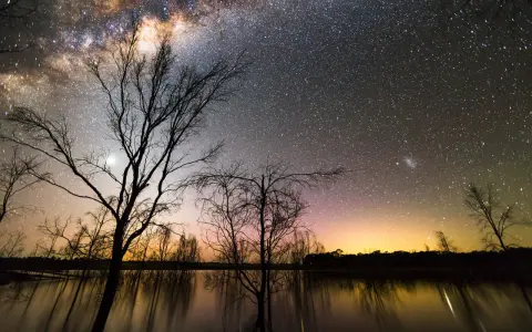 Canon EOS 2000D photo de nuit : une voie lactée spectaculaire au-dessus d'un lac reflétant les arbres dénudés et un ciel orangé.
