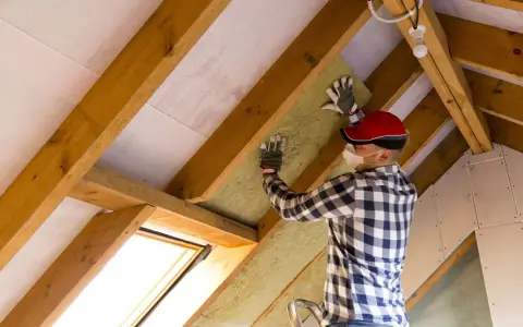 Uomo con mascherina e guanti installa materiale isolante tra le travi di legno di un sottotetto. È chiaro che conviene isolare il sottotetto per migliorare il comfort.