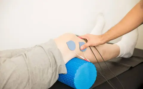 Rehabilitacja kolana w domu: elektrody na skórze, wałek do masażu pod nogą, ręka terapeuty.