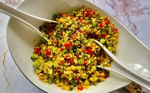 Kolorowa sałatka z kaszą bulgur, pomidorami, ogórkiem i ziołami, podana w białej misie z dwoma białymi szczypcami.