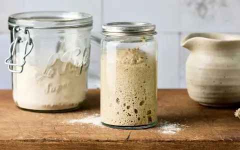 Blubbernder Sauerteig im Glas, bereit zum Backen. Daneben ein Glas mit Mehl, das zeigt, wie man den Sauerteig pflegen kann.