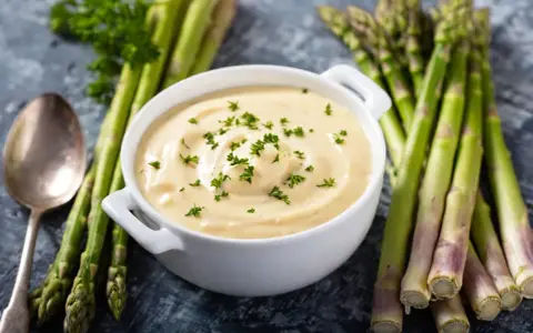 Un bol de sauce mousseline onctueuse, garni de persil frais, est présenté avec des asperges vertes et une cuillère.
