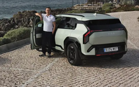 Mężczyzna opiera się o drzwi nowego, jasnoszarego Kia EV3, zaparkowanego na brukowanej drodze nad morzem.