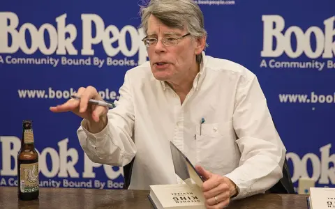 Stephen King, autore di libri da brivido, firma un volume. Sul tavolo, una bottiglia di root beer.