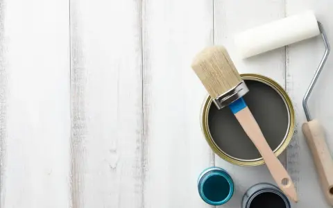 Herramientas para pintar mesitas de noche: lata de pintura gris, pincel, rodillo y botes de pintura azul y negra sobre fondo de madera blanca.