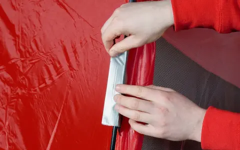 Hände reparieren ein rotes Zelt mit Klebeband.