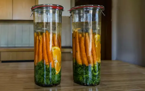 Zwei Gläser mit Karotten, Kräutern und Orangenscheiben in Salzlake zum Fermentieren.