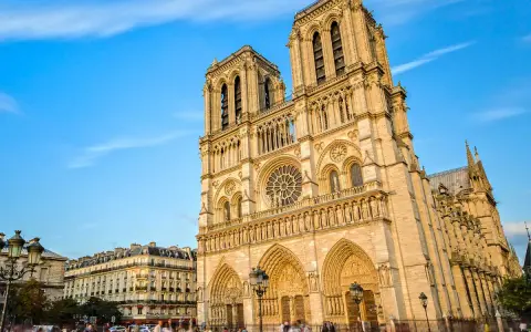 La majestueuse façade de Notre Dame de Paris se dresse sous un ciel bleu, ornée de détails gothiques. Des passants flânent sur la place.