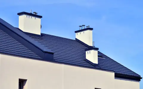 Nowoczesny dach bezokapowy z ciemnogranatową dachówką i białymi kominami, pod błękitnym niebem.
