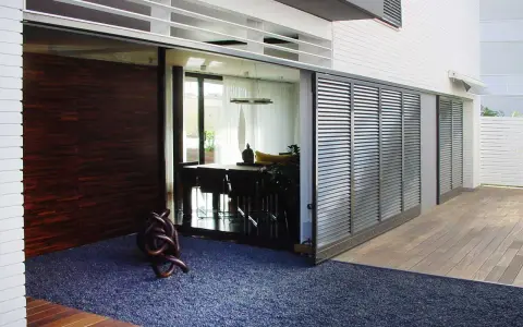 Una moderna casa con una contraventana corredera de lamas metálicas que protege un comedor interior.