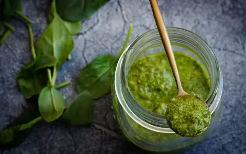 Słoik z zielonym pesto, na łyżce porcja sosu. Czy pesto ze słoika się podgrzewa? Świeże liście bazylii obok.