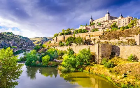 El **río Tajo** serpentea al pie de Toledo, una ciudad amurallada coronada por un imponente alcázar. Un puente de piedra cruza el agua, conectando las verdes orillas.