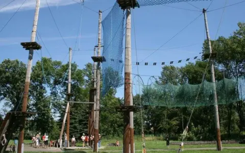 Śląski park linowy z drewnianymi słupami, siatkami i platformami na wysokości. Ludzie przygotowują się do zabawy.