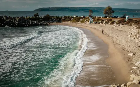 Piękne, turkusowe morze w Bułgarii obmywa piaszczystą plażę. W oddali widać wzgórza i samochody.