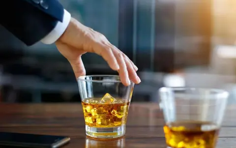 Hand greift nach einem Glas goldbraunen Whiskey mit Eiswürfeln. Ein Genuss, auch für Menschen, die glutenfreien Whiskey bevorzugen.