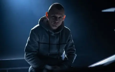 Młody mężczyzna w szarej bluzie z kapturem, z ogoloną głową, w mrocznym świetle. Jego spojrzenie sugeruje opowieść o polskim rapie.