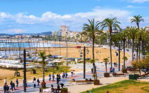 En livlig strandpromenad i Sitges, Spanien, med palmer, en sandstrand, båtar i hamnen och människor som njuter av solen.