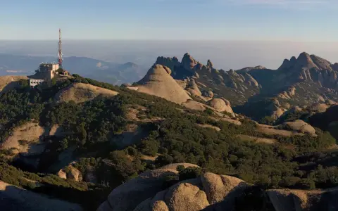 Edificio con antena en la cima de una montaña rocosa, con vistas a un paisaje montañoso y brumoso que se extiende hasta el horizonte, evocando la sensación de estar en planos cerca de Barcelona.