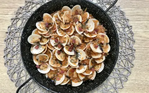 Almejas con jamón en una paellera, con trocitos de jamón y perejil picado, sobre un mantel de madera.