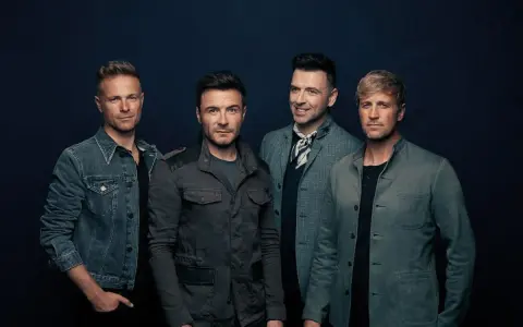 Medlemmar av Westlife poserar för en studiofotografering. De bär moderna jackor och ser direkt in i kameran.