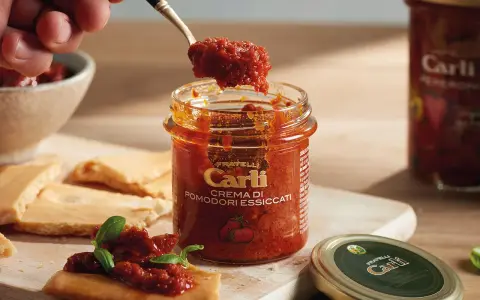 Une cuillère prélève de la crème de tomates séchées d'un pot Carli. Servie sur des crackers avec du basilic.