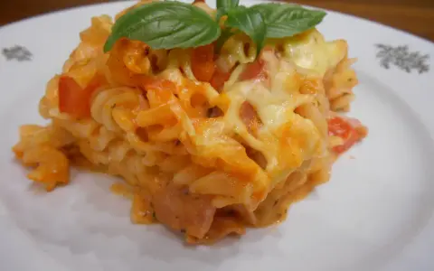 Pyszna zapiekanka z szynką, makaronem fusilli, pomidorami i ciągnącym się serem, ozdobiona listkami bazylii.
