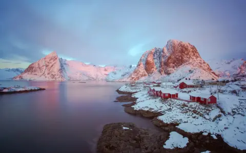 Vackert lofoten vinterlandskap med snöklädda berg, röda hus vid vattnet och en rosa himmel.