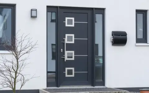 Nowoczesne drzwi zewnętrzne w ciemnoszarym kolorze z geometrycznymi przeszkleniami i metalowymi elementami. Obok skrzynka na listy.