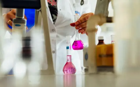 Ręce w białym fartuchu trzymające kolby z różowym płynem w laboratorium.