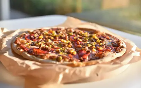 Apetyczna pizza Giuseppe 4 sery, z kukurydzą i mięsem, na papierze do pieczenia, w słonecznym świetle.