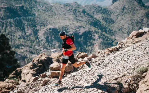 Un corredor de trail desciende por un sendero rocoso y empinado, con montañas boscosas al fondo.