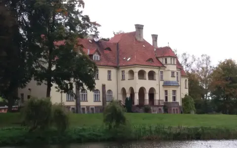 Malowniczy pałac z czerwoną, dachówkową, spadzistą dachem, otoczony zielenią i wodą.
