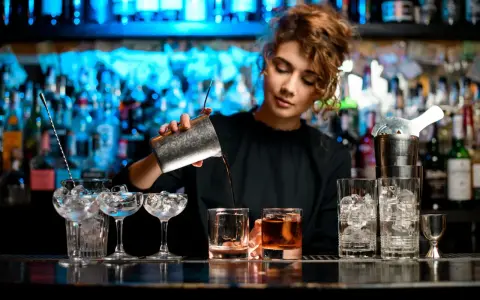 Barman nalewa drink z shakera do szklanki, w tle półki z butelkami i niebieskie światła.