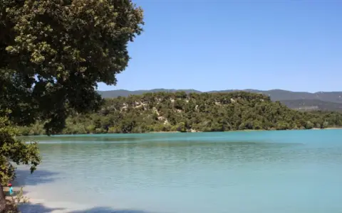 Vue paisible de l'étang de la Bonde, avec ses eaux turquoise bordées de verdure et de collines boisées sous un ciel bleu clair.