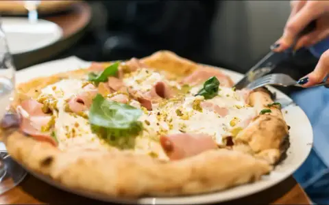 En person skär i en pizza bianco med skinka och basilika. Ett glas vatten står bredvid. Perfekt vin till pizza bianco.
