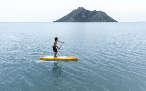 Chłopak na desce sup Aqua Marina przemierza spokojne morze w kierunku skalistej wyspy.