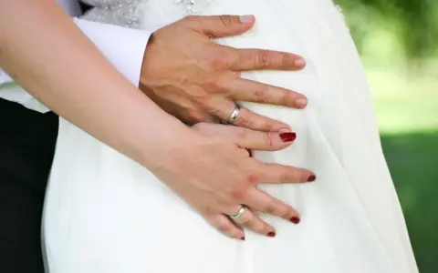 Hände auf dem Bauch einer schwangeren Braut im weißen Kleid. Ein Paar heiratet voller Vorfreude.