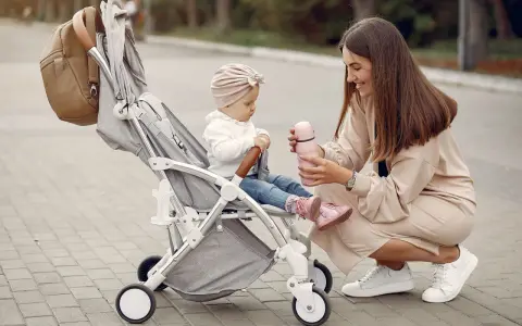 Mama karmi córeczkę z butelki, siedząc obok wózka. Idealne wózki dla dziewczynki na spacer.
