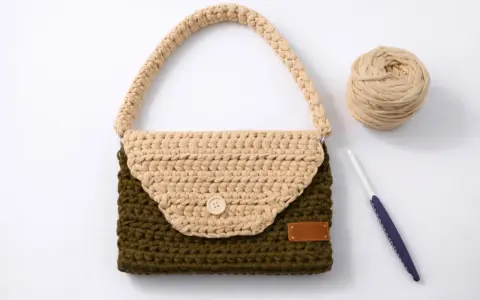 Bolso de trapillo bicolor, verde y beige, con asa y botón de madera. A su lado, ovillo de lana y ganchillo.